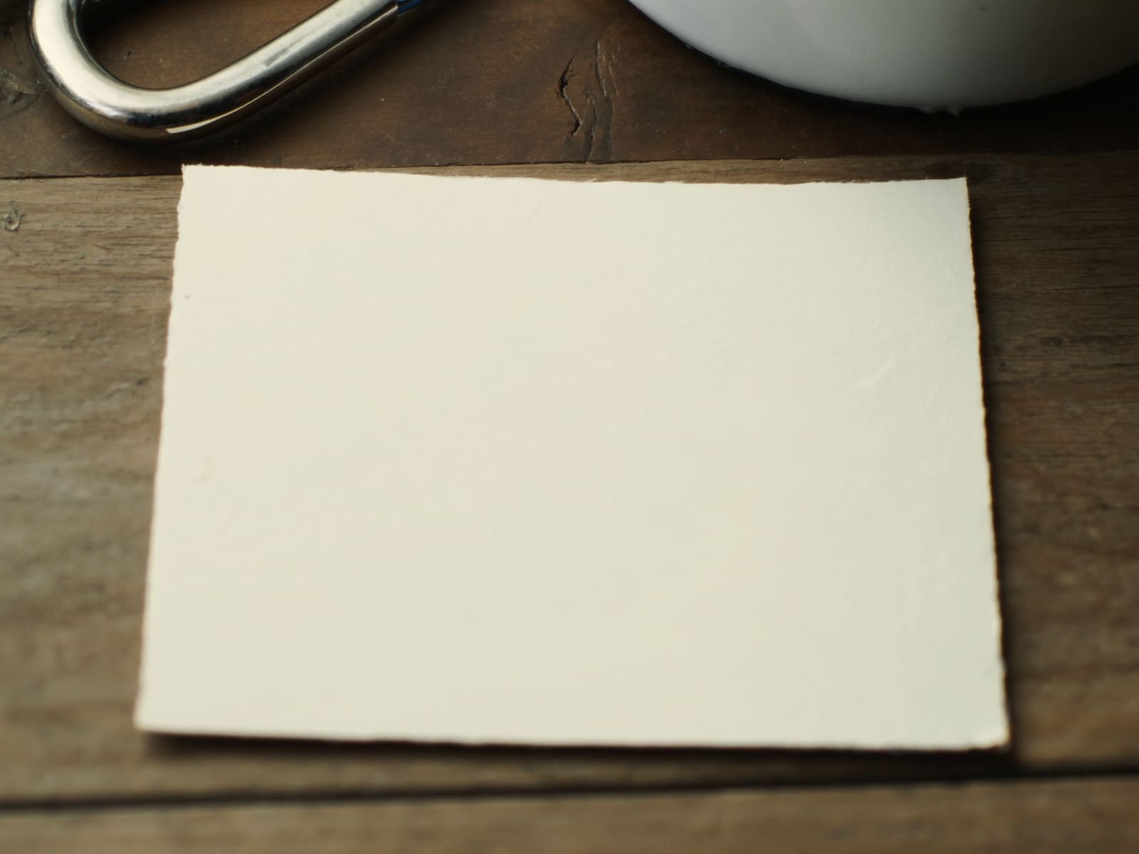 Unbedruckte Gutscheinkarte aus Büttenpapier auf einem Holztisch, daneben ein Karabiner und ein Emaillebecher.